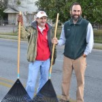 Two men standing on the side of a road holding brooms.
